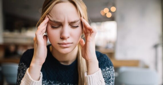Untitled-2 (4) Woman with closed eyes holding her temples with both hands, sitting indoors and appearing to experience discomfort or tension.