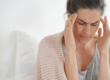 Woman sitting on a bed with her eyes closed and fingers pressing on her temples, showing discomfort or pain from a headache, stress, or tension possibly addressed by physical therapy at BioMotion Physical Therapy.