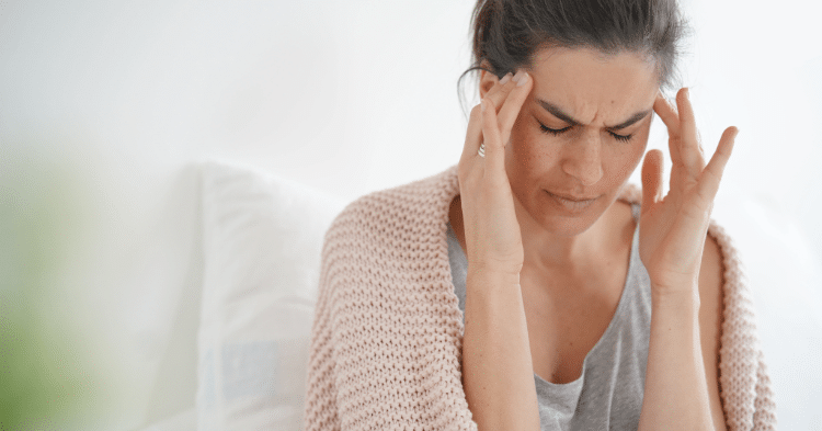 Woman sitting on a bed with her eyes closed and fingers pressing on her temples, showing discomfort or pain from a headache, stress, or tension possibly addressed by physical therapy at BioMotion Physical Therapy.