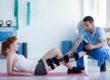 A woman with a knee brace lying on a yoga mat being assisted by a physical therapist in a gym setting.