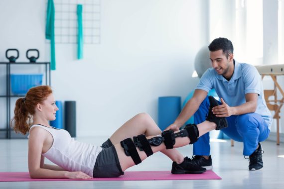 A woman with a knee brace lying on a yoga mat being assisted by a physical therapist in a gym setting.