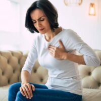cardiac-sq Woman sitting on a couch wearing a white shirt and blue jeans, holding her chest with a pained expression, suggesting discomfort or possible chest pain.