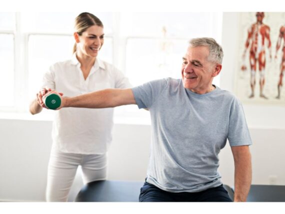 An older man sitting and lifting a green dumbbell with his arm extended, smiling as a female physical therapist in white supports and encourages him during a rehabilitation session.