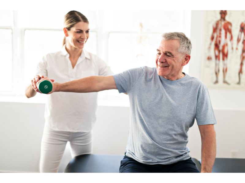 physical therapist helping a patient lift dumbells An older man sitting and lifting a green dumbbell with his arm extended, smiling as a female physical therapist in white supports and encourages him during a rehabilitation session.