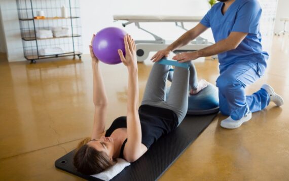 AdobeStock_423996099 A woman lying on a mat holding a purple exercise ball above her chest while a physical therapist in blue scrubs assists her with a leg resistance band exercise in a rehab or therapy center.