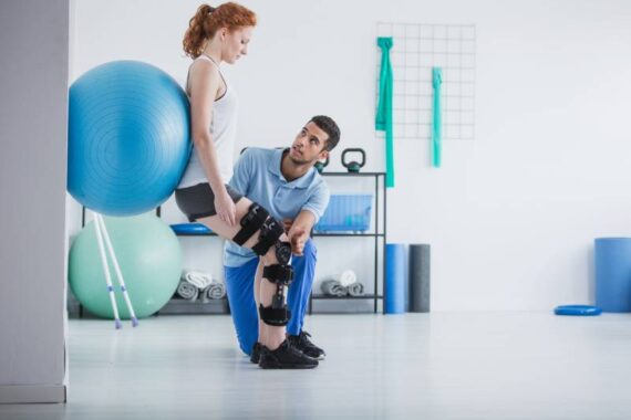 Female physical therapy patient in a leg brace balancing a blue exercise ball against the wall with her back as her physical therapist stabilizes her injured leg.