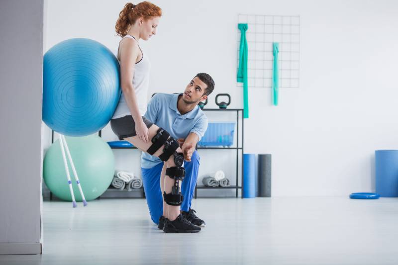 How Long Is Physical Therapy? Female physical therapy patient in a leg brace balancing a blue exercise ball against the wall with her back as her physical therapist stabilizes her injured leg.