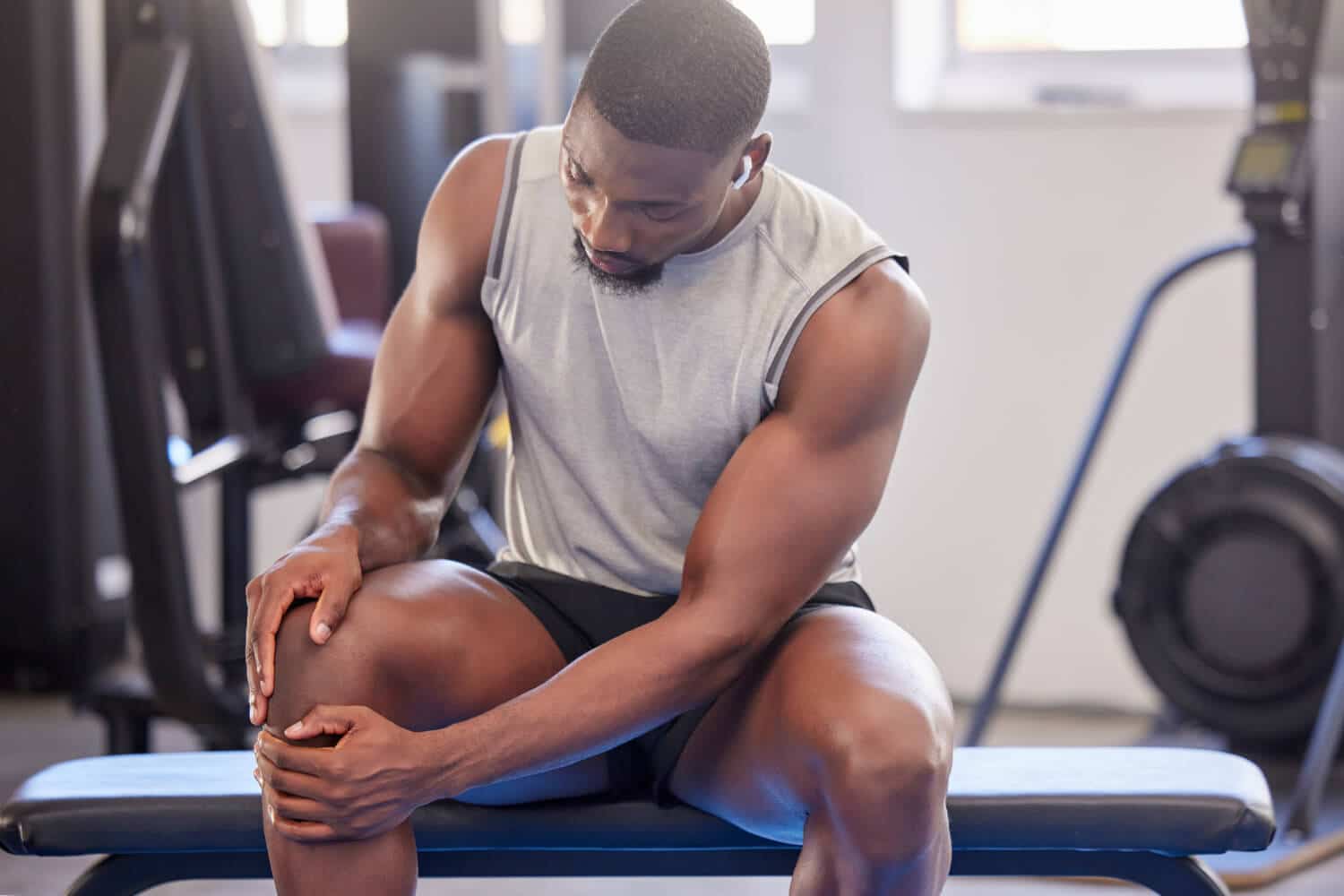 knee-pain-in-gym Man in athletic wear sitting on a bench at the gym, holding his knee with both hands and looking down at it.