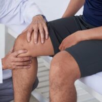 knee-pain-treatment Close-up of a physical therapist examining a patient's knee, with both individuals seated, focusing on the patient's bent leg.