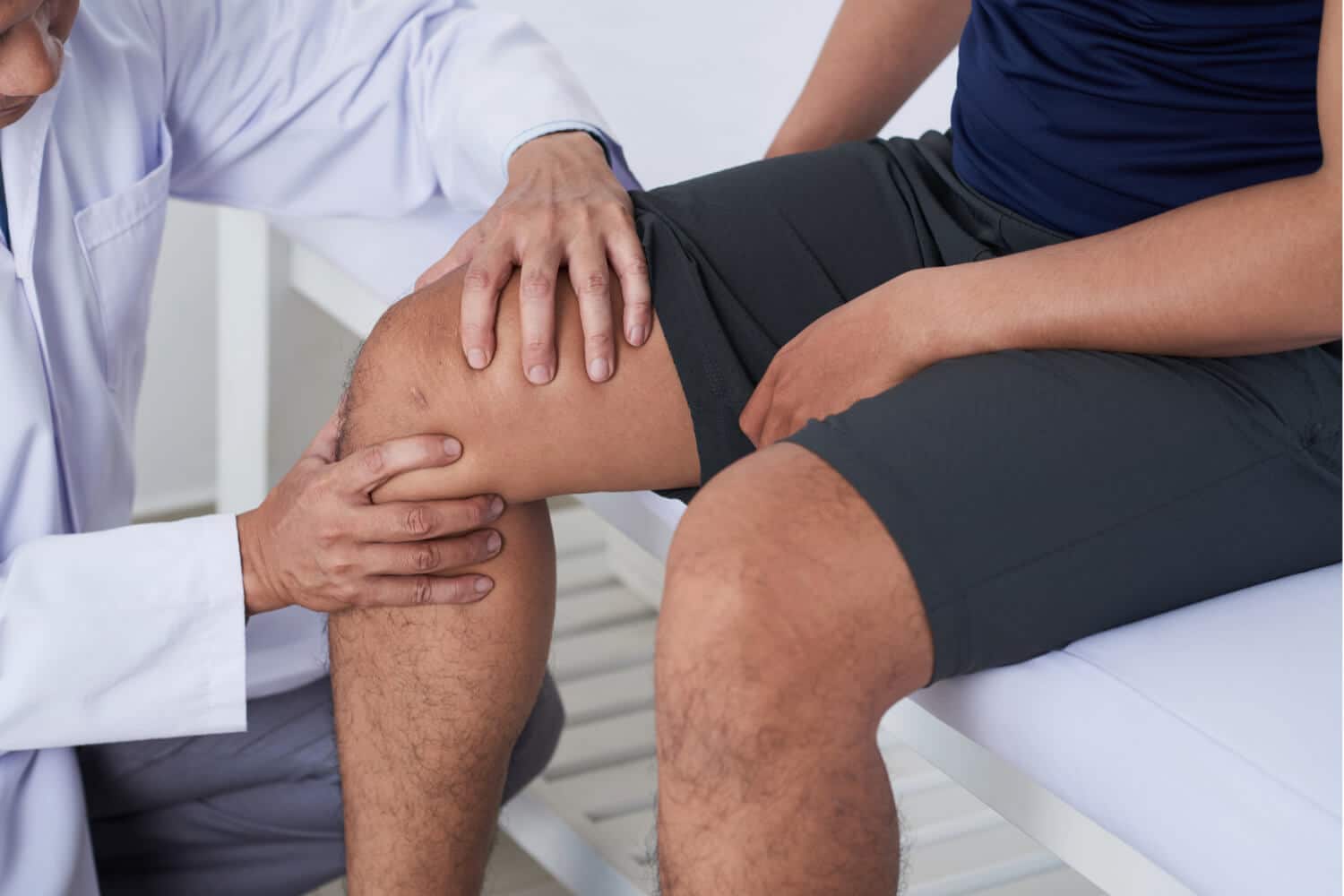 knee-pain-treatment Close-up of a physical therapist examining a patient's knee, with both individuals seated, focusing on the patient's bent leg.