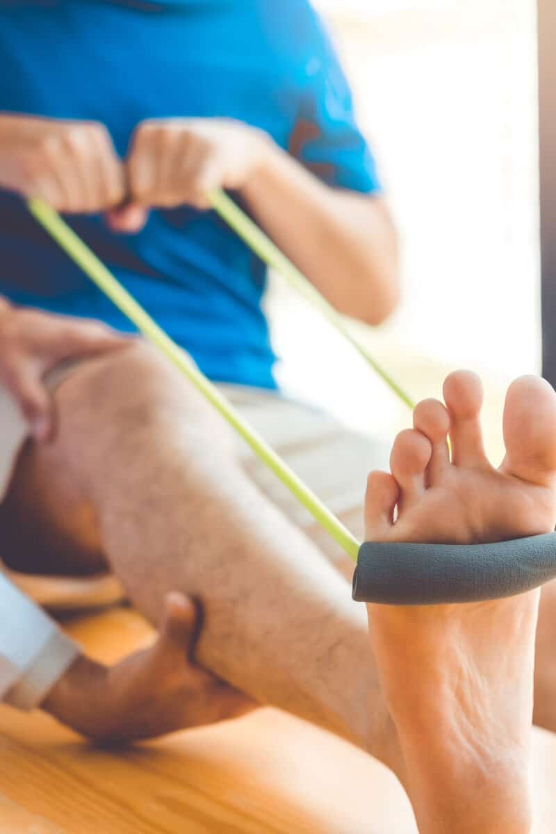 knee-recovery Close-up of a person using a green resistance band looped around their foot while another individual supports their leg, suggesting a therapeutic or rehabilitation exercise.