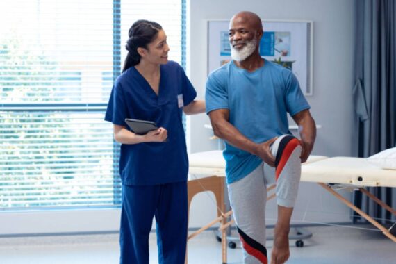 Young female physiotherapist helps older Black male patient move his legs.
