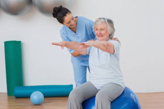Physiotherapist helps an elderly white woman work on her balance by using an exercise ball.