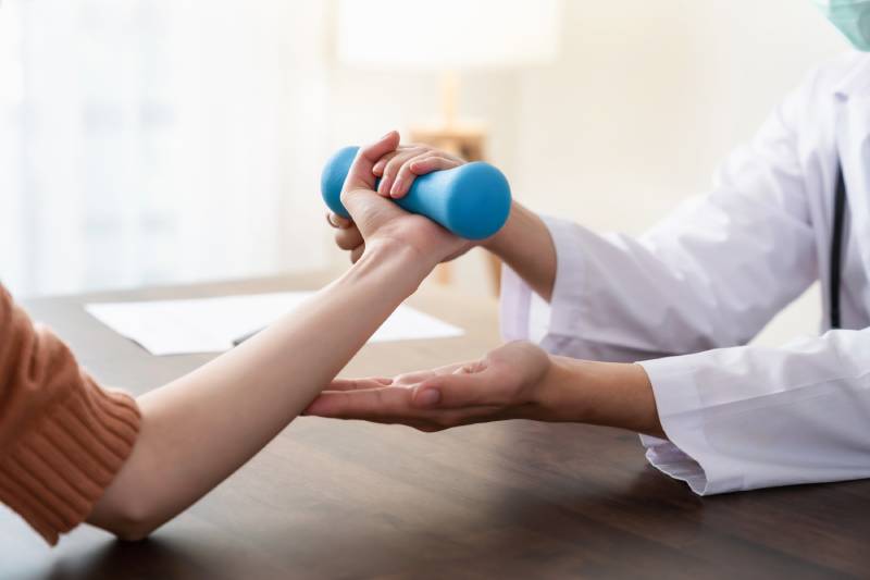 Physical therapist helps female patient work on strength with light dumbbell exercise.