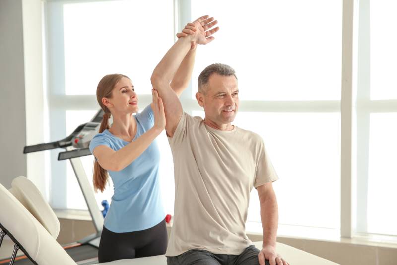 A physical therapist assists a man in a light t-shirt with an overhead arm stretch while he sits on a treatment table in a brightly lit clinic.