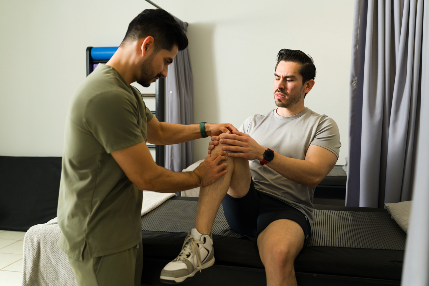 A physical therapist examines a seated man's knee in a clinic setting.