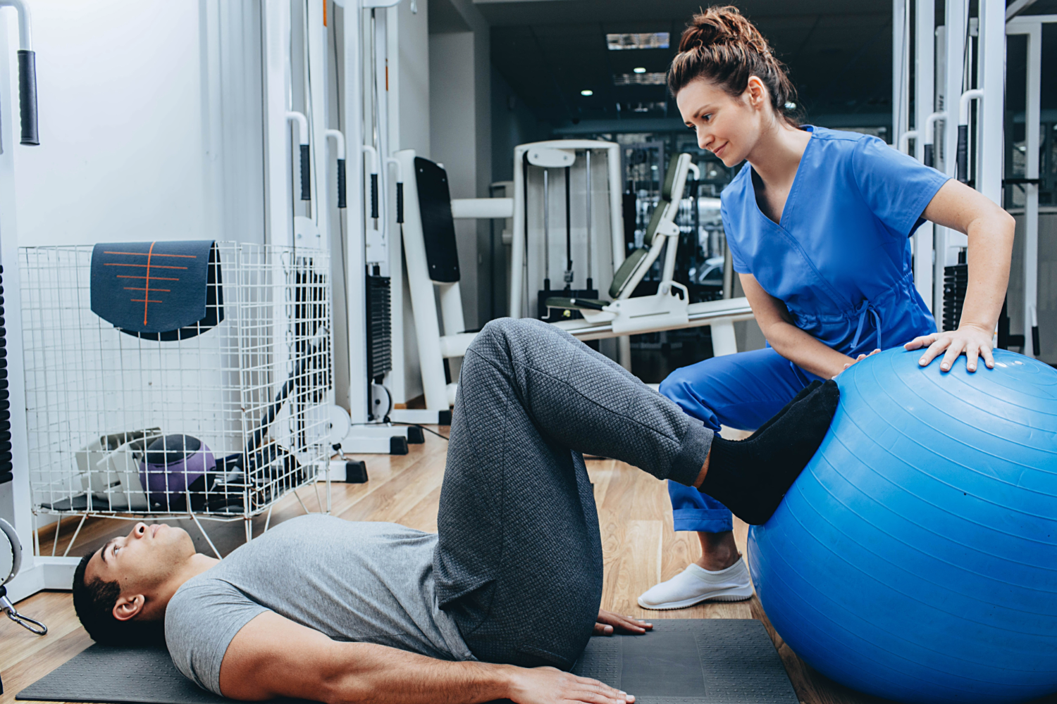 Female physical therapist helps a male patient with leg exercises including a blue exercise ball.