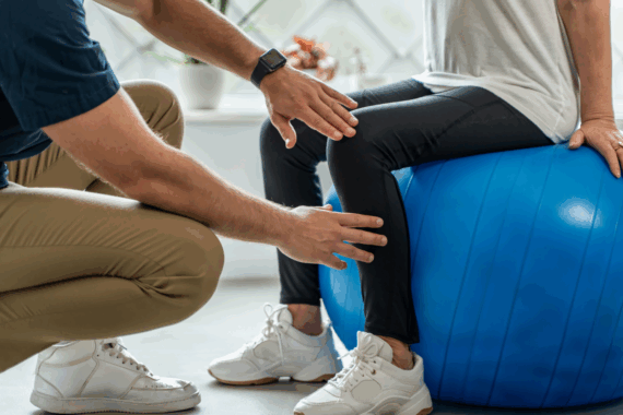Physiotherapist helping senior woman with leg rehabilitation exercise on balance ball.