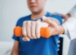 A young man holds out an orange weight at chest level with the assistance of a doctor. He is sitting down in a chair.