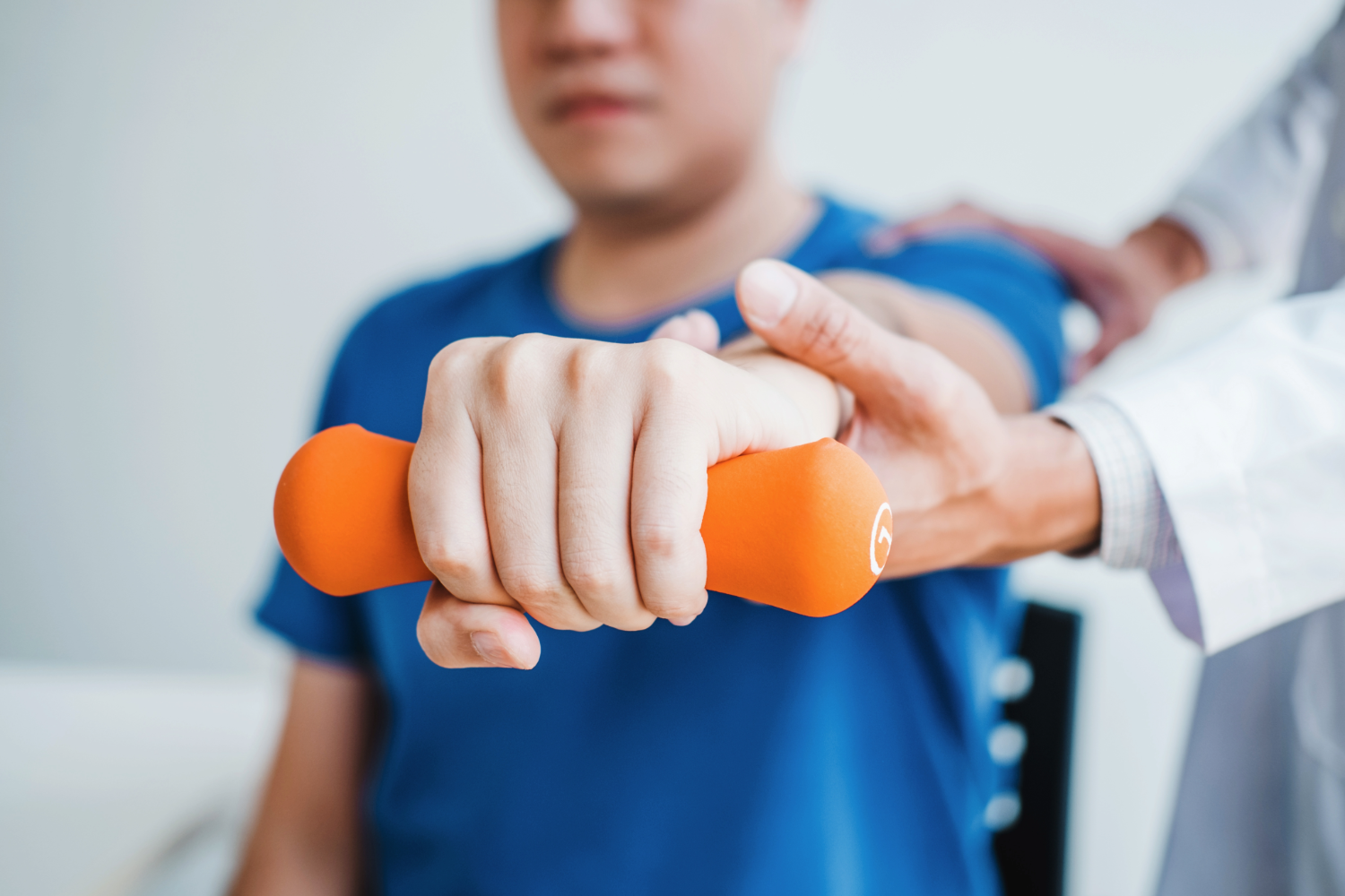 A young man holds out an orange weight at chest level with the assistance of a doctor. He is sitting down in a chair.