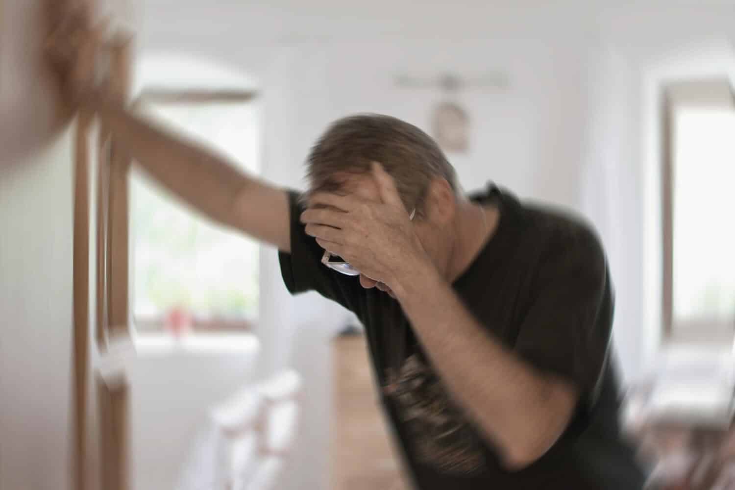 Blurred image of a man appearing dizzy or disoriented, holding his head with one hand and steadying himself against a wall with the other, suggesting symptoms related to a balance disorder.