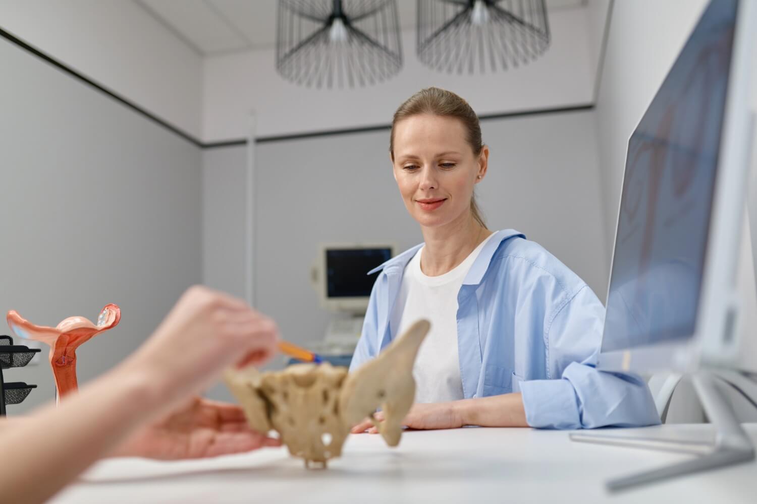 Woman in a medical office attentively listening as a professional uses a pelvic bone model to explain pelvic floor anatomy, with a uterus model and medical equipment visible nearby.