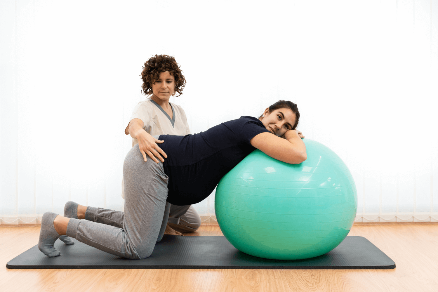 Pregnant woman on hands and knees leaning over an exercise ball while a therapist provides support and guidance, demonstrating a pelvic floor rehabilitation exercise in a clinical setting.
