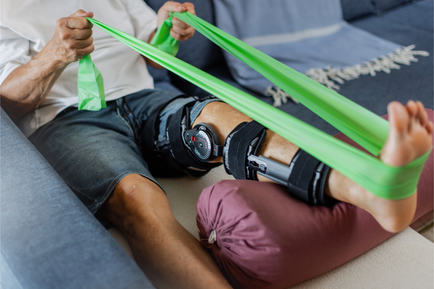Senior man performing rehabilitation exercises using a green resistance band to aid in his knee recovery process at home. A physical therapy exercise with a leg brace.
