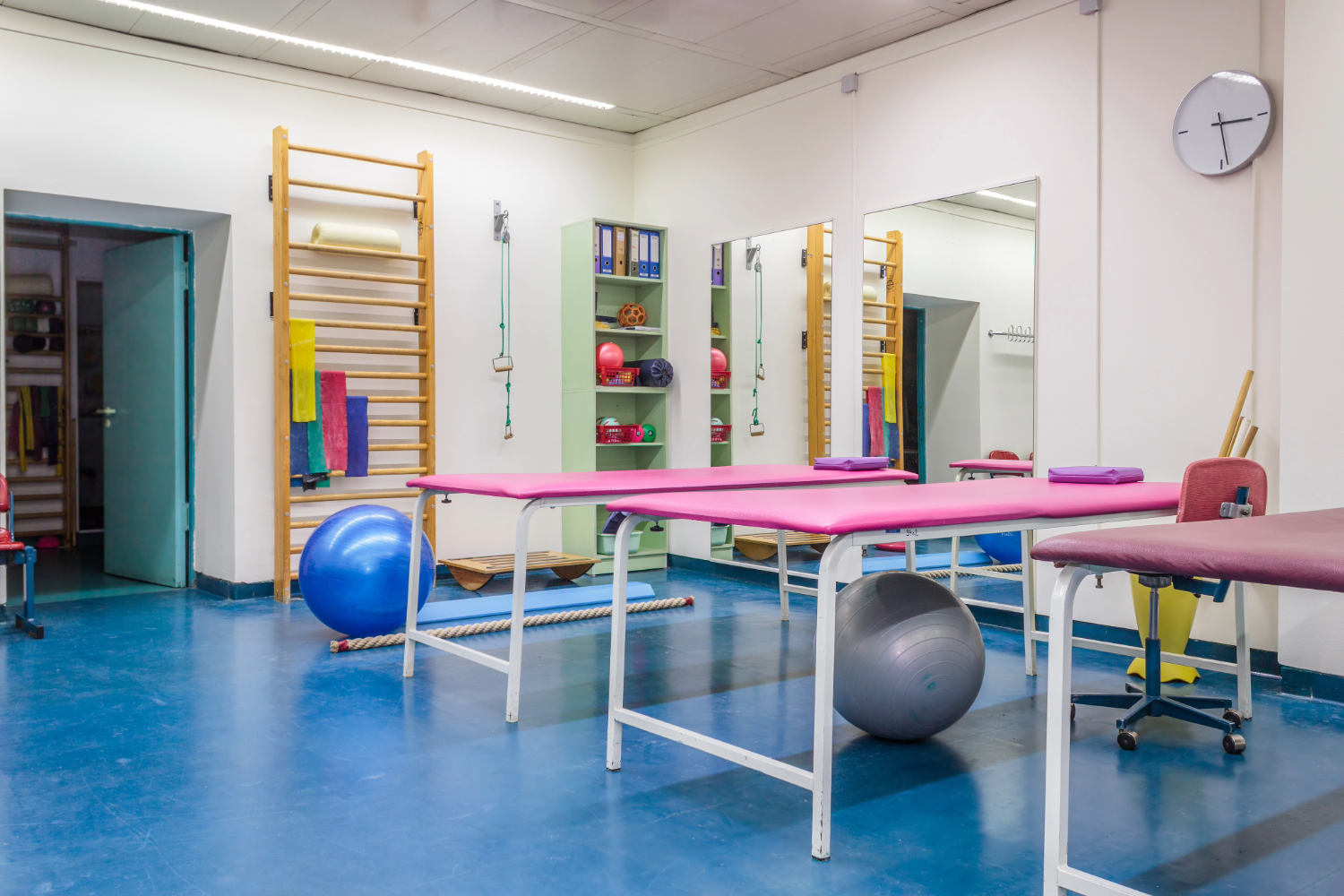 Empty room in physiotherapy clinic. Tables line the room with exercise balls underneath, and two shelves of smaller physical therapy equipment.