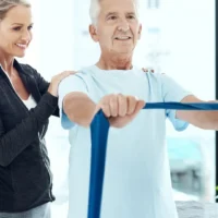 elder man stretching and exercise band out in front of him with woman standing behind him smiling and holding his shoulders