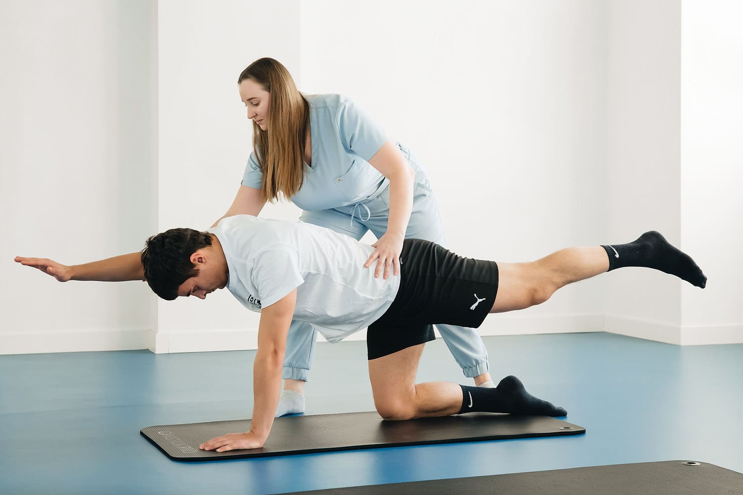 back-pain-pt-supervised-core-exercise Physical therapist supervising a patient performing a bird-dog core exercise on a mat in a clinic.