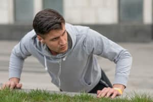 Man in a gray hoodie doing outdoor push-ups beside a grassy area.