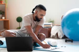 Man sitting cross-legged on an exercise mat taking notes on a clipboard during a virtual physical therapy consultation on his laptop.